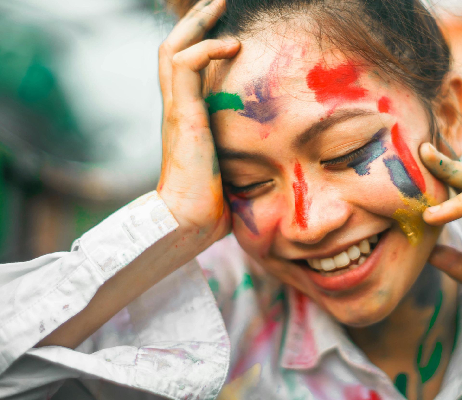 Cheerful young ethnic female with dye on face touching forehead and cheeks with eyes closed on blurred background