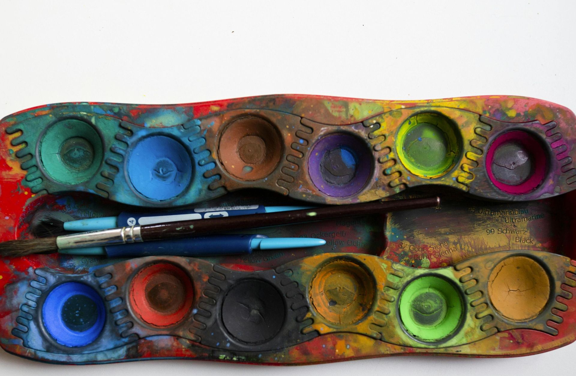 A colorful watercolor palette with various paint wells and brushes on a white surface.